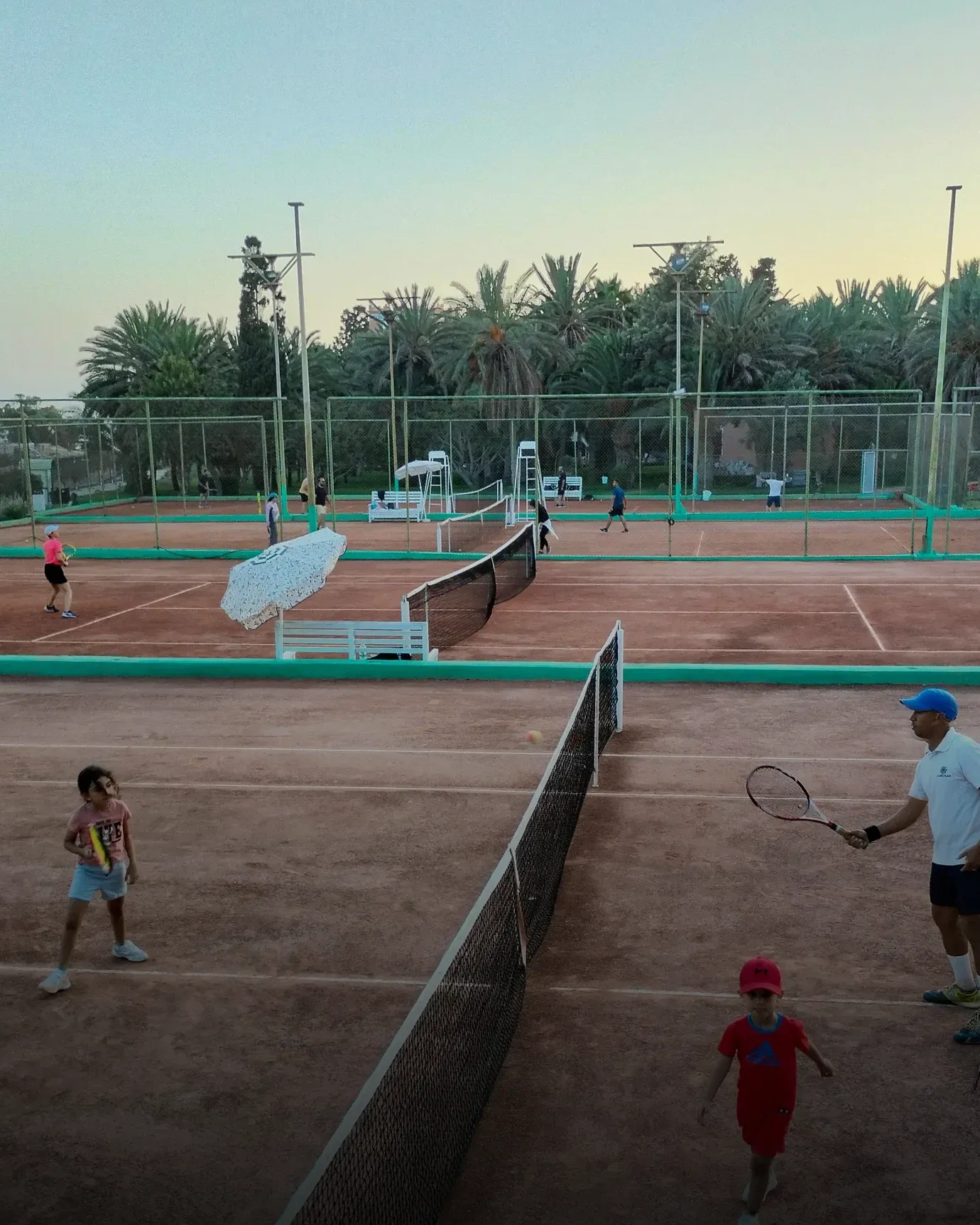 Scuola di tennis per giovani: bambini in allenamento con il loro maestro, rete in primo piano, a fine giornata