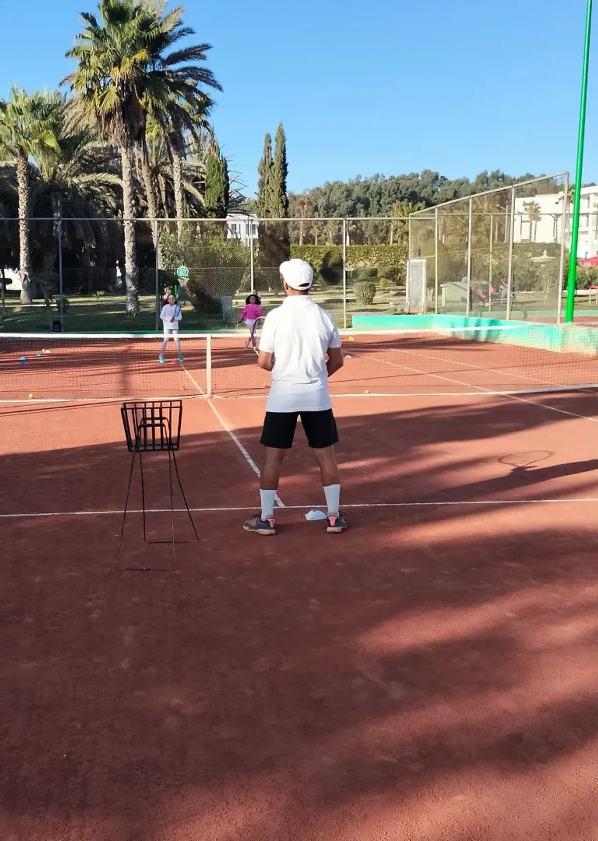 Tennisstunde im Club in Agadir: ein Trainer am Ballkorb, seine Spieler auf der anderen Seite des Netzes, in voller Sonne