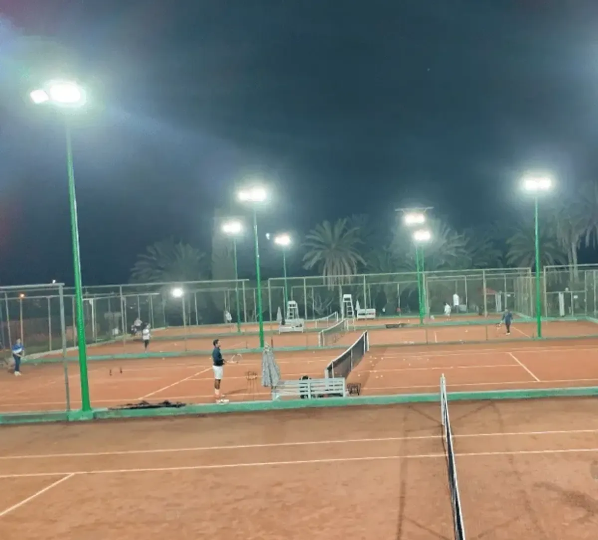 Les courts en terre battue du club éclairés en soirée, joueurs à l’échange