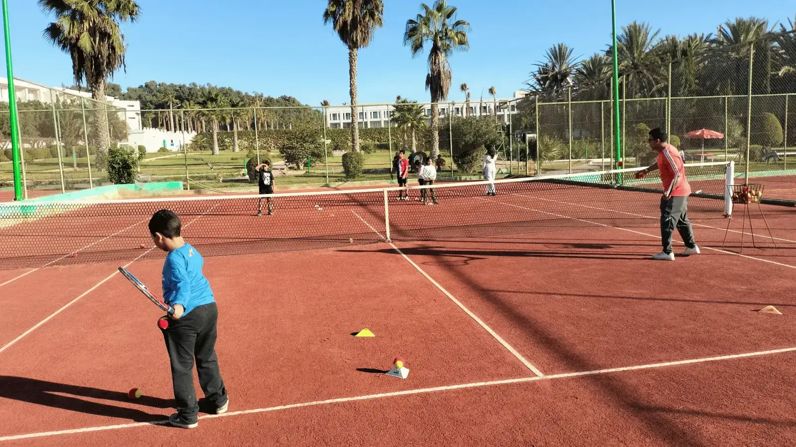 Mini Tennis à Agadir : un garçon en bleu, raquette et balle rouge en main, et un coach qui lance des balles depuis son panier